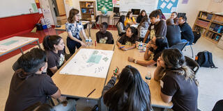 Students sit around a table working on a big paper activity while receiving feedback from an educator.