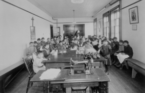  Children in a classroom with a sewing machine on a table. A nun stands in the back of the classroom. Taken circa 1929.