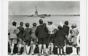 A group of children in 1930s era clothing stare and point at the Statue of Liberty.