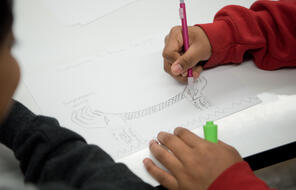 Close view of a middle school student in a red sweatshirt writing on a piece of paper with a pencil.