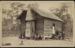 Photo shows a log cabin with two African American men seated outside and an African American woman standing in the doorway of a slave or sharecropper dwelling.