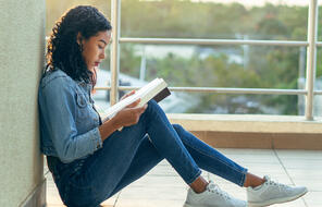 A girl leans against a wall while reading a book.