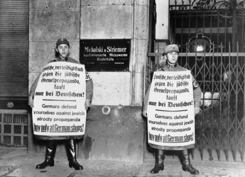 SA members in 1933 stand in front of a barricaded Jewish shop, holding signs in both German and English urging the boycott of Jewish businesses.