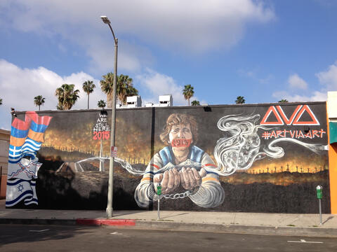 A mural features an elderly woman with a piece of tape over her mouth with "1915" written on it.