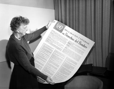 A black and white photo of Eleanor Roosevelt holding a large copy of the Universal Declaration of Human Rights in Spanish.