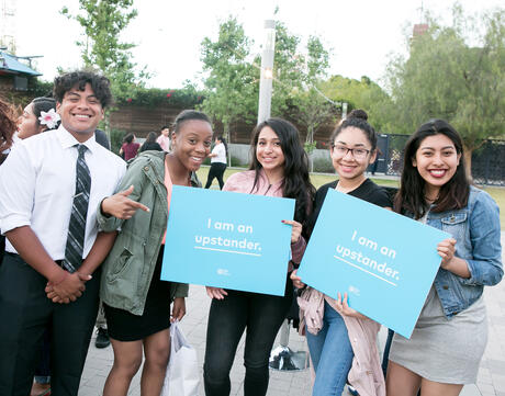 Five high school students smiling and two of them holding blue signs that say "I am an Upstander"